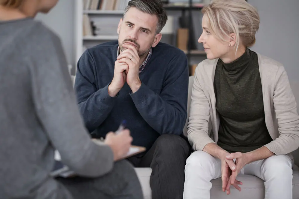 La moglie sostiene il marito in terapia con l'uomo che ascolta incuriosito il counselor in un ufficio grigio